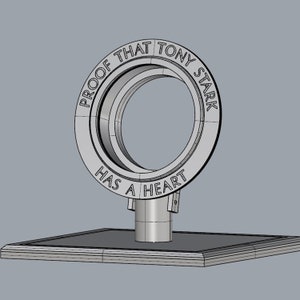 May include: A silver metal ring with the words "Proof that Tony Stark has a heart" engraved around it. The ring is on a square base.