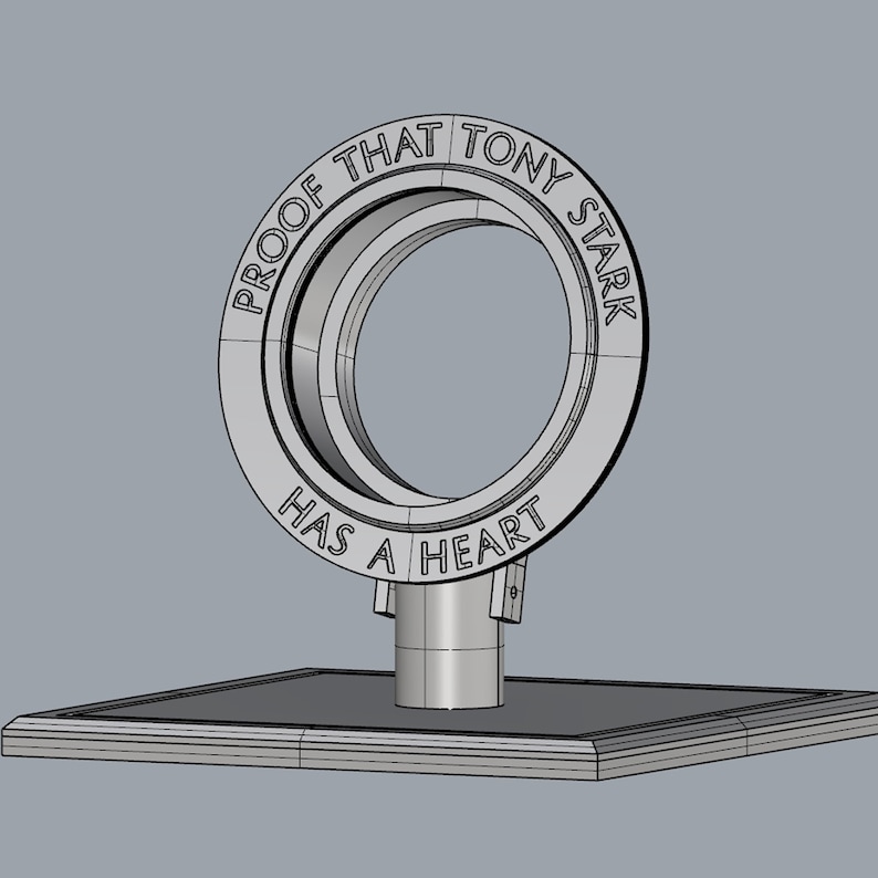 May include: A silver metal ring with the words "Proof that Tony Stark has a heart" engraved around it. The ring is on a square base.