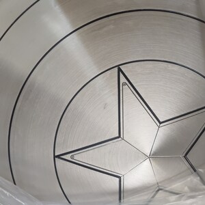 May include: A silver metal Captain America shield with a black five-pointed star in the center. The shield has concentric circles around the star.
