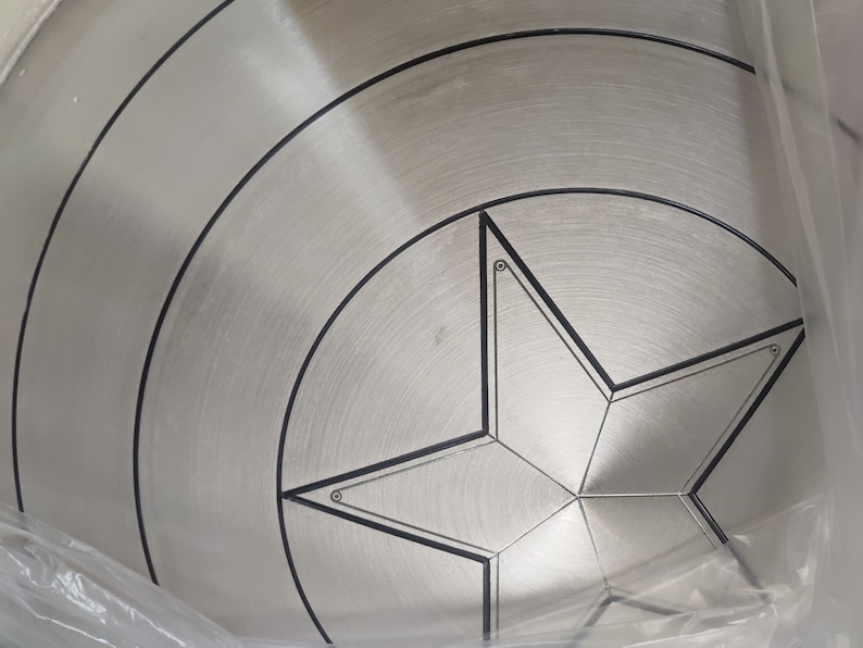 May include: A silver metal Captain America shield with a black five-pointed star in the center. The shield has concentric circles around the star.