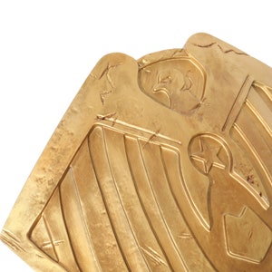 May include: A gold-colored shield with a stylized eagle and star design. The shield has a distressed, aged look with visible scratches.