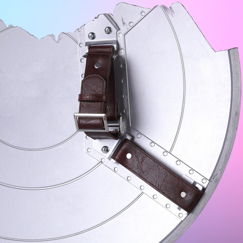May include: A silver-colored shield with a distressed edge, featuring brown leather straps and a metal buckle. The shield has concentric circular grooves and is constructed with metal plates.