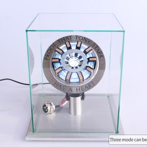 Puede incluir: Una réplica del reactor de arco de Iron Man de color plateado y cobre con una luz azul, expuesta en una vitrina de vidrio. La inscripción dice "Proof that Tony Stark has a heart".