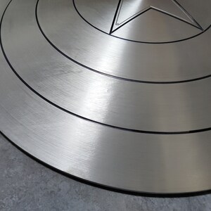 May include: A silver-colored Captain America shield with a red, white, and blue star in the center. The shield has a brushed metal finish and concentric circles around the star.