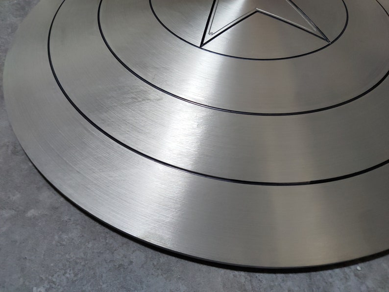 May include: A silver-colored Captain America shield with a red, white, and blue star in the center. The shield has a brushed metal finish and concentric circles around the star.
