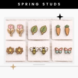 May include: Six pairs of wooden stud earrings displayed on white cards, featuring designs of pink tulips, green leaves, orange carrots, brown flowers, yellow and black bees, and pink and blue butterflies. The text SPRING STUDS appears at the top.