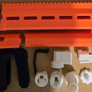Puede incluir: Un conjunto de piezas de plástico naranja y blanco para un juguete de pistola. Las piezas incluyen una culata, un cañón, un guardamanos y un cargador. Las piezas son todas de plástico y tienen un acabado liso.