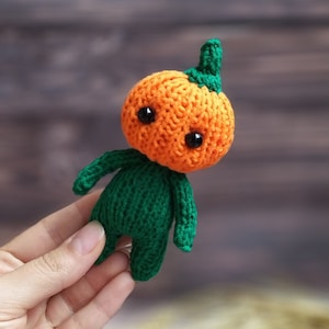 May include: A small crocheted pumpkin-shaped toy with a green body and an orange head. The toy has two black eyes and a green stem on top of its head.