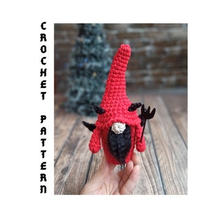 May include: A handmade red crochet gnome with black horns and a black beard, holding a black trident. The gnome has a long, pointed hat and a white mustache. The text "CROCHET PATTERN" is on the left side.