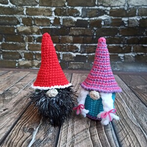 May include: Two crocheted gnomes, one with a red hat and a black beard, and the other with a pink and purple hat and a white beard.