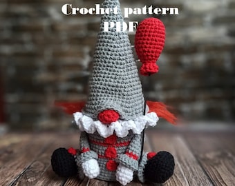 May include: Crocheted clown gnome with a red balloon, wearing a gray and white hat, a white ruffle collar, and a red and white striped outfit. The gnome is sitting on a wooden surface. Crochet pattern PDF.