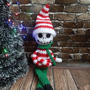 Puede incluir: Un mu&ntilde;eco de elfo de Navidad de crochet con una cara de calavera blanca, que lleva un gorro a rayas rojo y blanco y una bufanda verde. El elfo est&aacute; sentado en un suelo de madera.