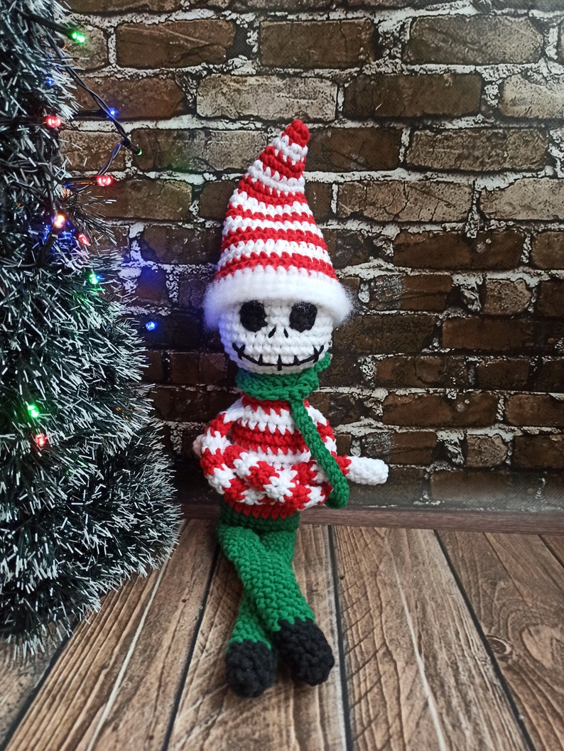 Puede incluir: Un mu&ntilde;eco de elfo de Navidad de crochet con una cara de calavera blanca, que lleva un gorro a rayas rojo y blanco y una bufanda verde. El elfo est&aacute; sentado en un suelo de madera.