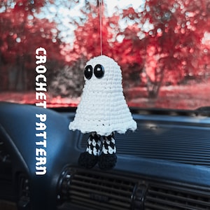 Puede incluir: Fantasma blanco de crochet con ojos de botón negros colgando de un espejo retrovisor en un coche. El fantasma tiene piernas rayadas en blanco y negro. CROCHET PATTERN está escrito verticalmente en el lado izquierdo de la imagen.