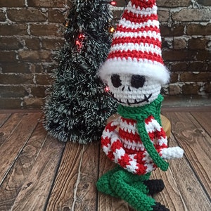 Puede incluir: Un mu&ntilde;eco de elfo de Navidad de crochet con un gorro a rayas rojas y blancas, pantalones verdes y una bufanda verde. El elfo tiene una cara blanca con ojos negros y una boca negra.