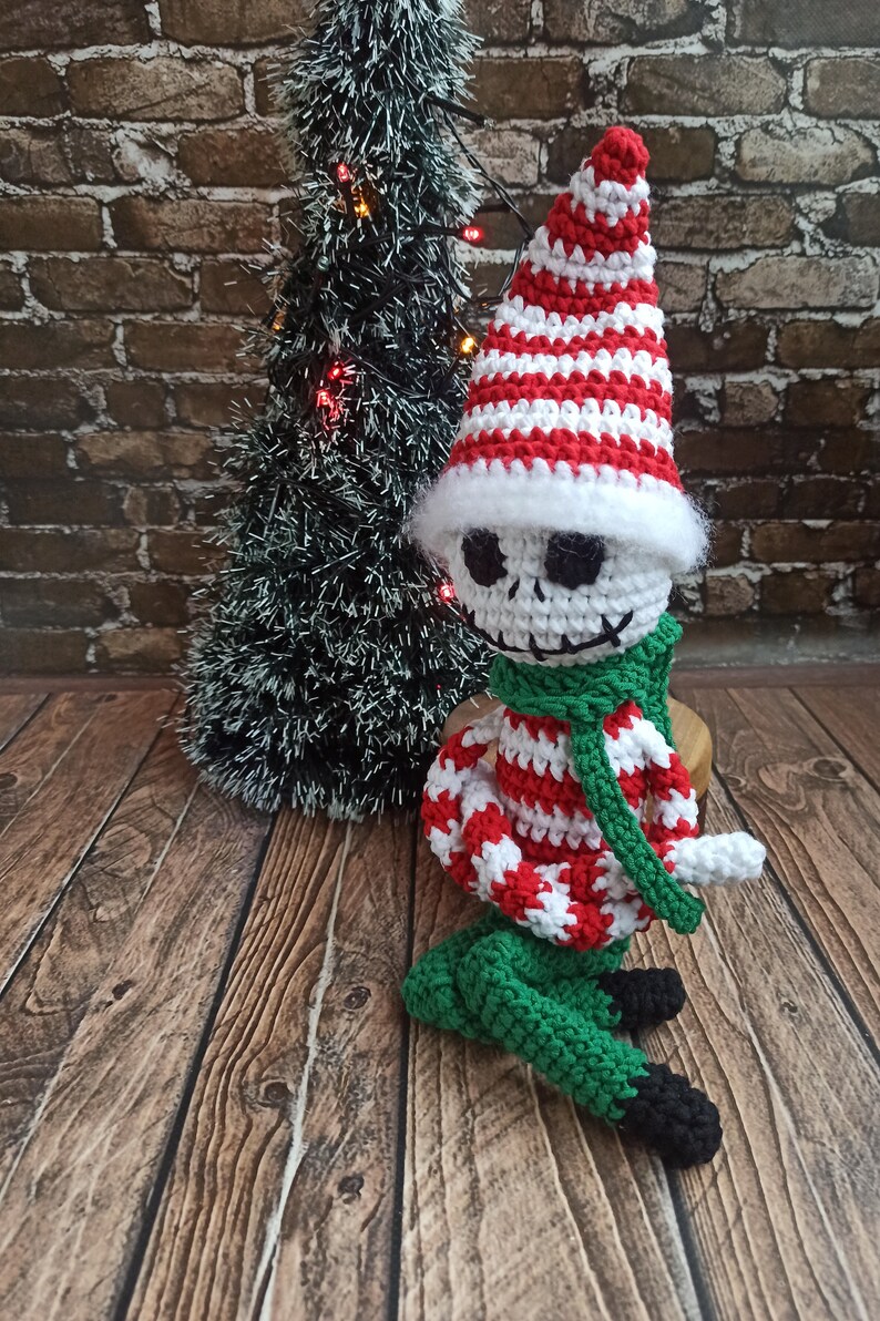 Puede incluir: Un mu&ntilde;eco de elfo de Navidad de crochet con un gorro a rayas rojas y blancas, pantalones verdes y una bufanda verde. El elfo tiene una cara blanca con ojos negros y una boca negra.