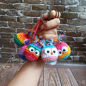 Pode incluir: Um conjunto de chaveiros de coruja de crochê coloridos. Cada coruja é de uma cor diferente e tem um padrão diferente. Os chaveiros são presos a um anel de metal.