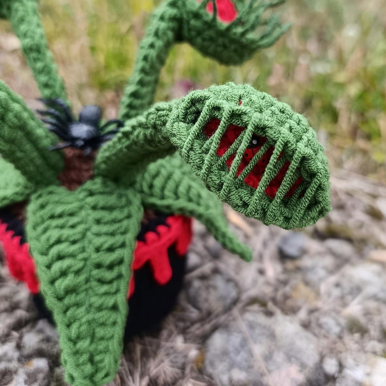 May include: A handmade crochet Venus flytrap plant with green leaves and red accents inside the traps. The plant is in a black pot with red details and a small black spider. The plant is a unique decorative item.