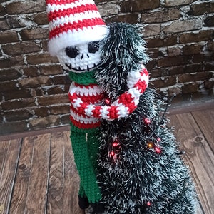 Puede incluir: Un mu&ntilde;eco de esqueleto de crochet con un gorro y un su&eacute;ter a rayas rojas y blancas, abrazando un &aacute;rbol de Navidad de crochet verde y blanco con luces rojas.