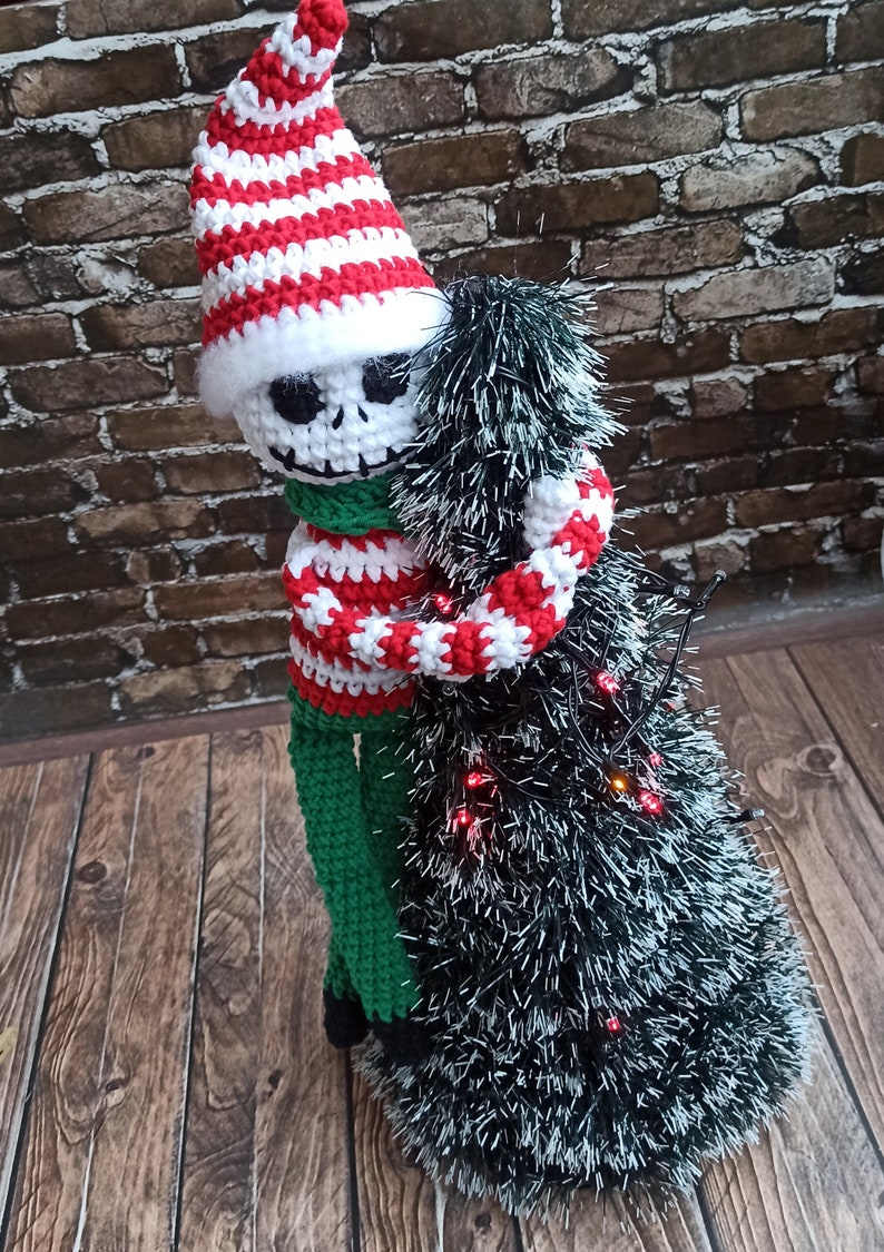 Puede incluir: Un mu&ntilde;eco de esqueleto de crochet con un gorro y un su&eacute;ter a rayas rojas y blancas, abrazando un &aacute;rbol de Navidad de crochet verde y blanco con luces rojas.