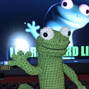 Può includere: Una rana verde all'uncinetto fatta a mano con occhi bianchi e neri e un'espressione sorridente. La rana è davanti a uno schermo di computer con un'immagine di rana dei cartoni animati. Il testo "Lizard Li" è visibile sullo schermo.