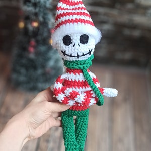 Puede incluir: Una mu&ntilde;eca de esqueleto de crochet con un gorro a rayas rojas y blancas, una bufanda verde y un su&eacute;ter a rayas rojas y blancas. La mu&ntilde;eca tiene pantalones verdes y zapatos negros.