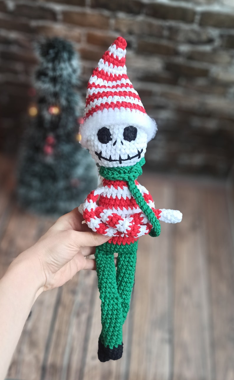 Puede incluir: Una mu&ntilde;eca de esqueleto de crochet con un gorro a rayas rojas y blancas, una bufanda verde y un su&eacute;ter a rayas rojas y blancas. La mu&ntilde;eca tiene pantalones verdes y zapatos negros.