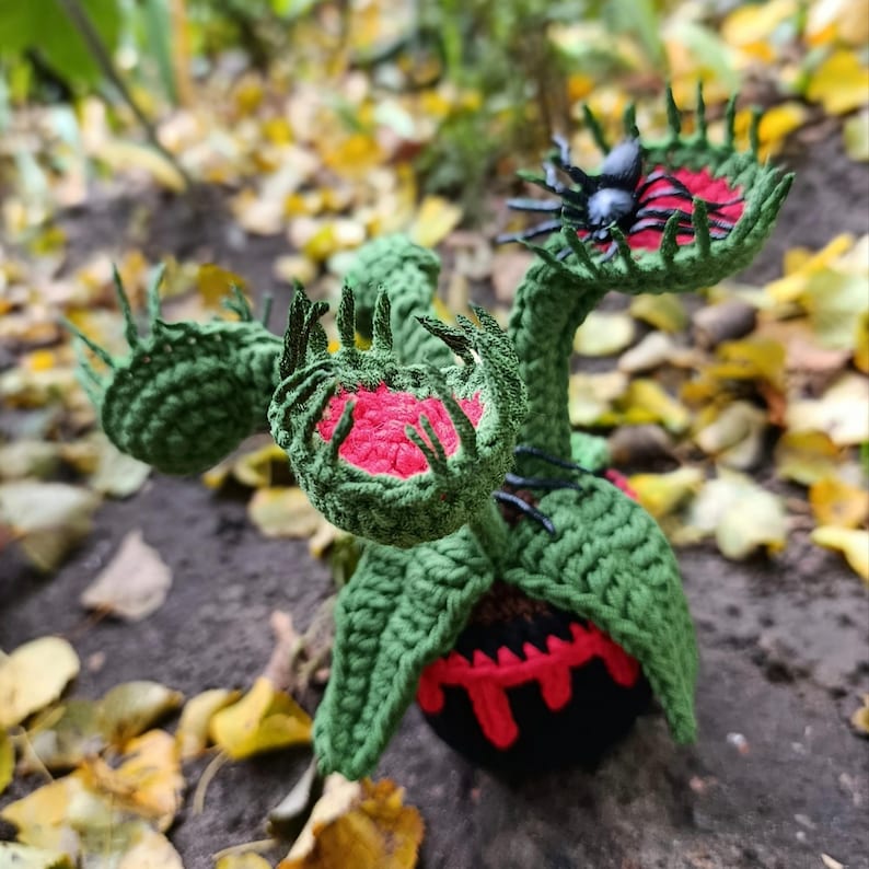 May include: A handmade, green crocheted Venus flytrap with red and black accents. The plant features open, tooth-edged traps with red interiors and a black base with red detailing. A black spider sits atop one trap.