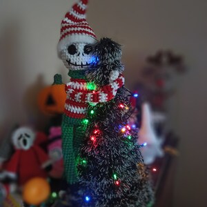 Puede incluir: Un esqueleto de crochet con un gorro a rayas rojas y blancas abraza un peque&ntilde;o &aacute;rbol de Navidad negro y peludo con luces de colores.