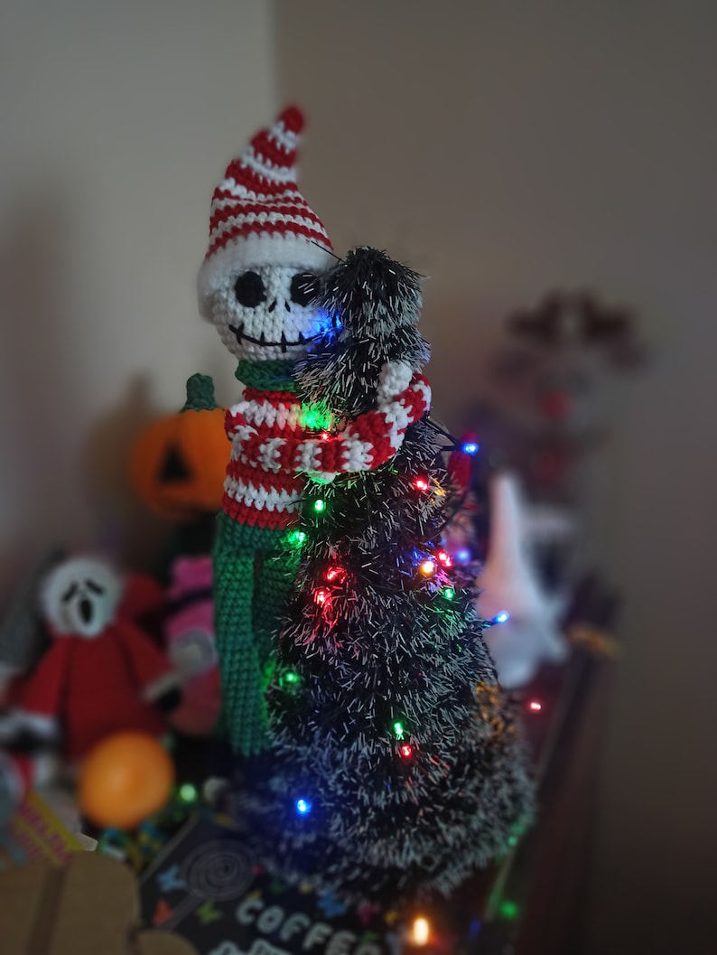 Puede incluir: Un esqueleto de crochet con un gorro a rayas rojas y blancas abraza un peque&ntilde;o &aacute;rbol de Navidad negro y peludo con luces de colores.