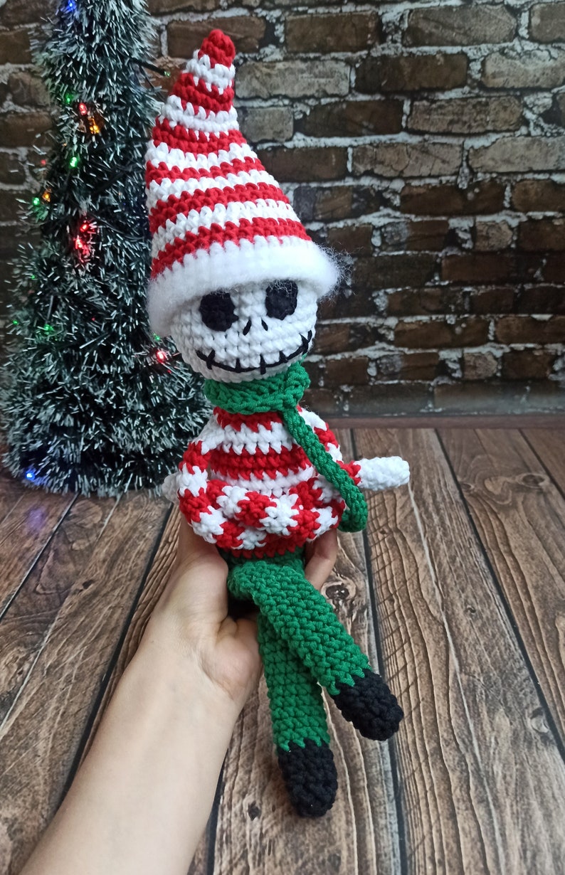Puede incluir: Un mu&ntilde;eco de elfo de Navidad de crochet con un gorro a rayas blanco y rojo, una bufanda verde y pantalones a rayas verdes y negros.