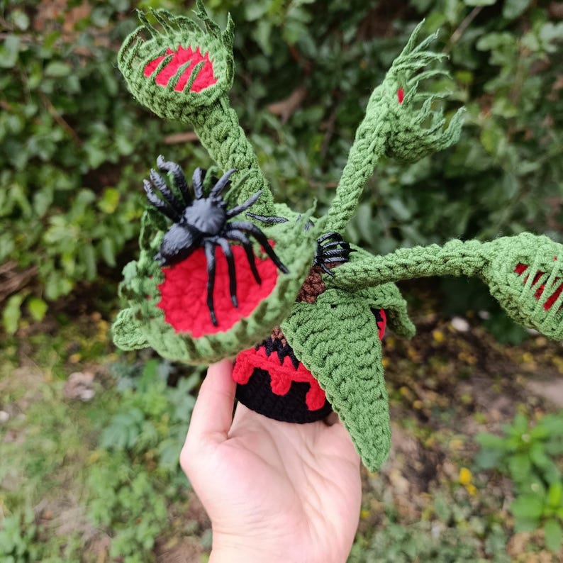 May include: A handmade crochet Venus flytrap plant with a black spider on one of the leaves. The plant is green with red accents and a black base. The plant is held in a hand.
