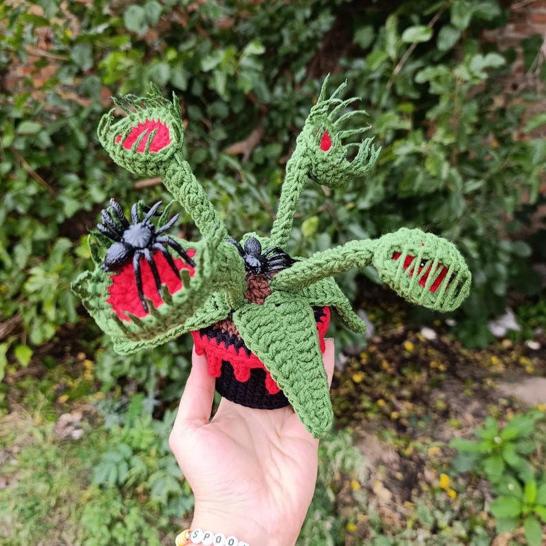 May include: Crocheted Venus flytrap plant with green leaves, red interiors, and black spiders. The plant is in a black and red pot. The handmade plant is a unique decorative item.