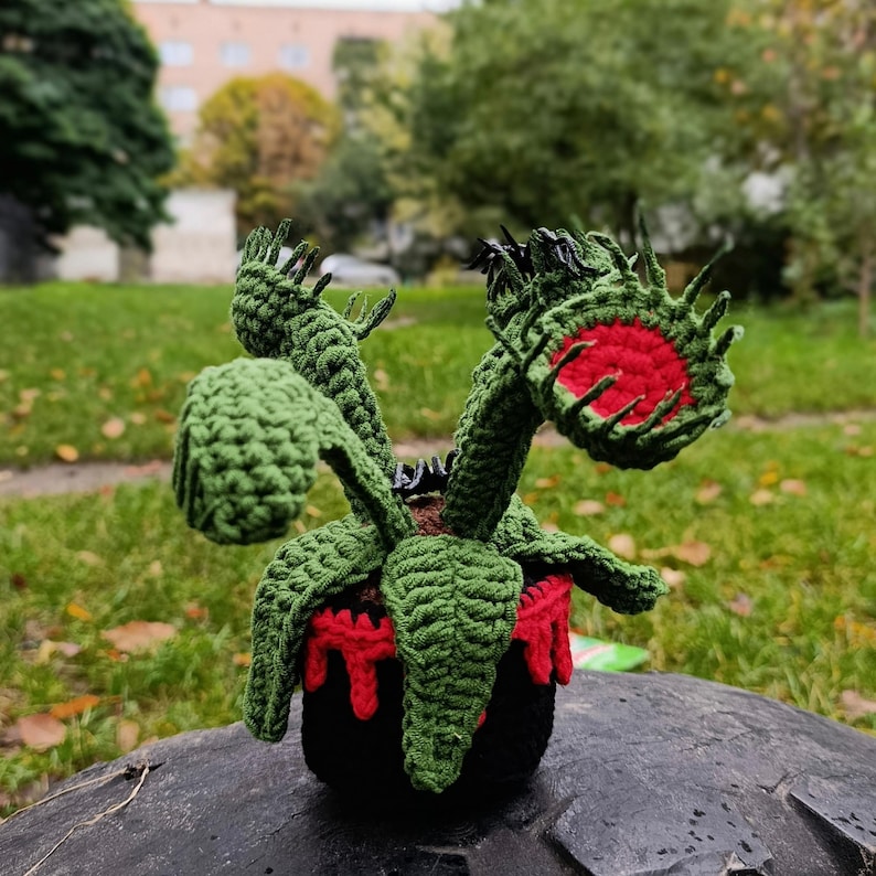 May include: A handmade crochet Venus flytrap plant. The plant features green leaves and red-lined traps, set in a black pot with red accents. The plant is a unique, whimsical decorative item.