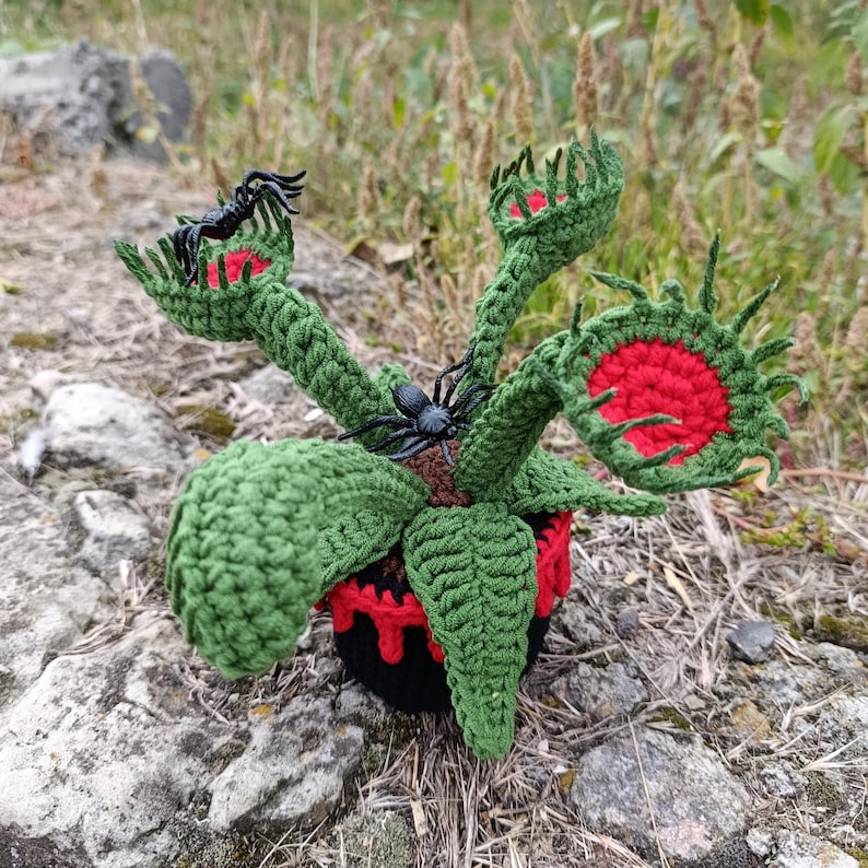 May include: A crocheted Venus flytrap plant with green leaves and red-lined traps. The plant is in a black pot with red accents and features two black plastic spiders. The plant is sitting on a rock.