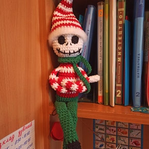 Puede incluir: Un mu&ntilde;eco de elfo de Navidad de crochet con un gorro a rayas blanco y rojo, una cara de calavera blanca, una bufanda verde y pantalones verdes. El mu&ntilde;eco est&aacute; sentado en una estanter&iacute;a con libros y una tabla de letras.