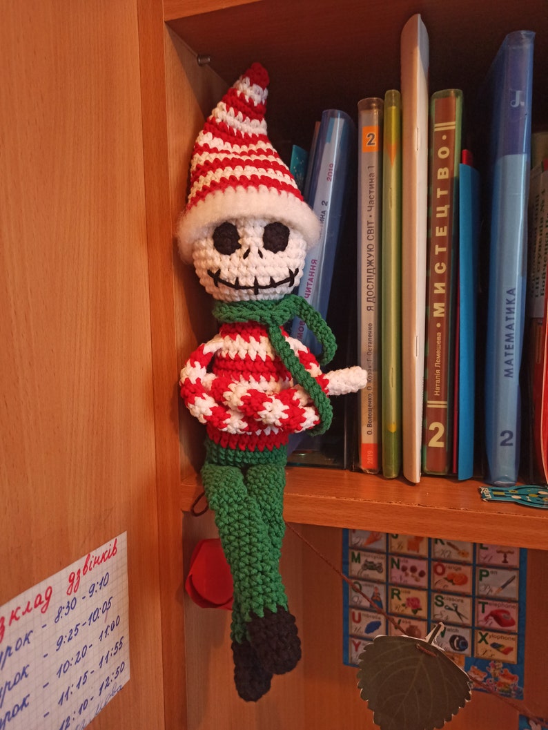 Puede incluir: Un mu&ntilde;eco de elfo de Navidad de crochet con un gorro a rayas blanco y rojo, una cara de calavera blanca, una bufanda verde y pantalones verdes. El mu&ntilde;eco est&aacute; sentado en una estanter&iacute;a con libros y una tabla de letras.