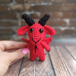 May include: A red crocheted goat with black horns and eyes. The goat is standing on a wooden surface.