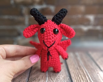 May include: A red crocheted goat with black horns and eyes. The goat is standing on a wooden surface.