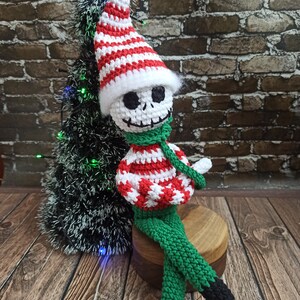 Puede incluir: Un mu&ntilde;eco de esqueleto de elfo de crochet con un gorro a rayas rojas y blancas y pantalones verdes. El mu&ntilde;eco est&aacute; sentado en un taburete de madera frente a un peque&ntilde;o &aacute;rbol de Navidad artificial.