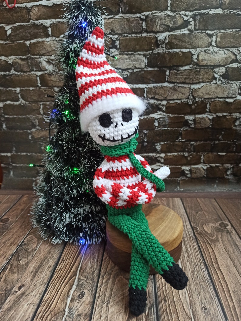 Puede incluir: Un mu&ntilde;eco de esqueleto de elfo de crochet con un gorro a rayas rojas y blancas y pantalones verdes. El mu&ntilde;eco est&aacute; sentado en un taburete de madera frente a un peque&ntilde;o &aacute;rbol de Navidad artificial.