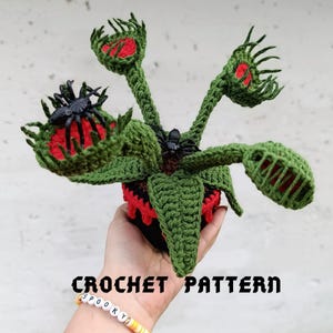 May include: A crocheted Venus flytrap plant with green leaves, red interiors, and black spiders. The plant is in a black and red pot. The text "CROCHET PATTERN" is displayed below the plant. A bracelet with the word "SPOOKY" is visible.