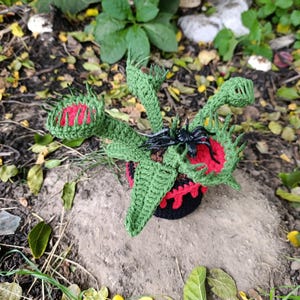 May include: A handmade crochet Venus flytrap plant with green leaves and red and green traps. A black plastic spider sits on one of the traps. The plant is in a black and red pot, set on a rock.
