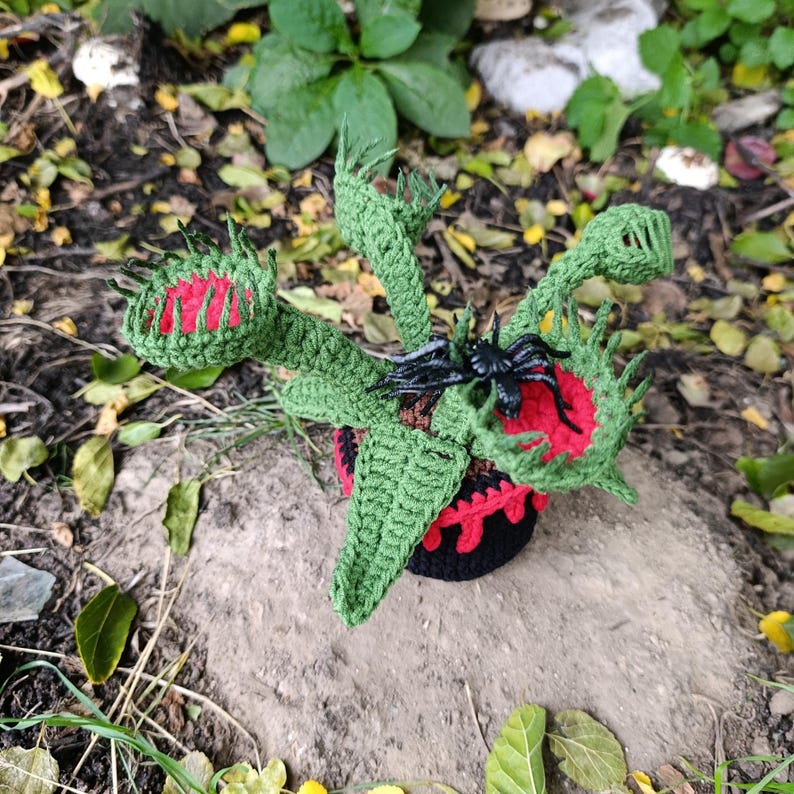 May include: A handmade crochet Venus flytrap plant with green leaves and red and green traps. A black plastic spider sits on one of the traps. The plant is in a black and red pot, set on a rock.