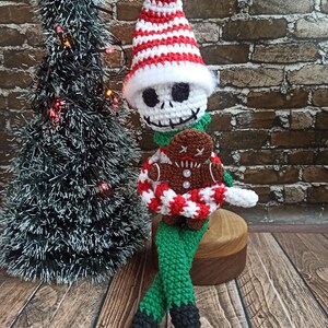 Puede incluir: Un mu&ntilde;eco de elfo de Navidad de crochet con un gorro a rayas blanco y rojo, pantalones verdes y zapatos negros. El elfo sostiene una galleta de jengibre.