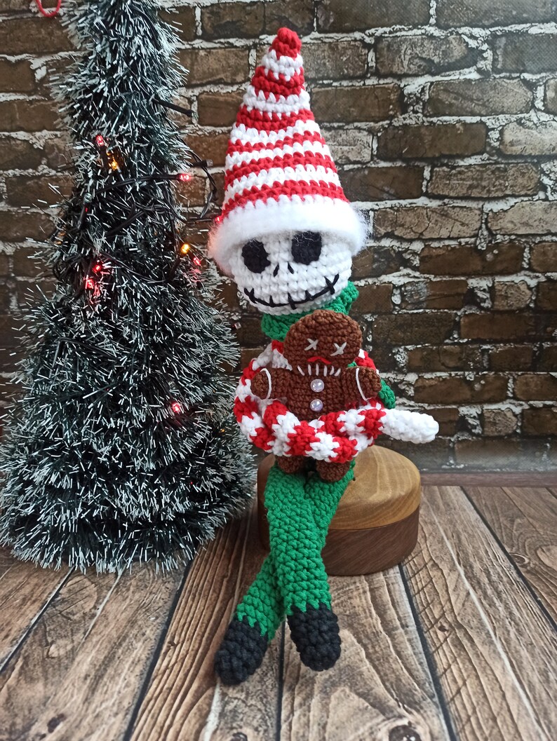 Puede incluir: Un mu&ntilde;eco de elfo de Navidad de crochet con un gorro a rayas blanco y rojo, pantalones verdes y zapatos negros. El elfo sostiene una galleta de jengibre.