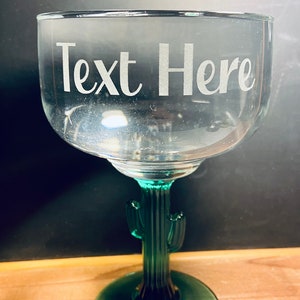 May include: A clear margarita glass with a green cactus-shaped stem and base. The glass has the words "Text Here" etched on the bowl. The glass is designed for cocktails and beverages.