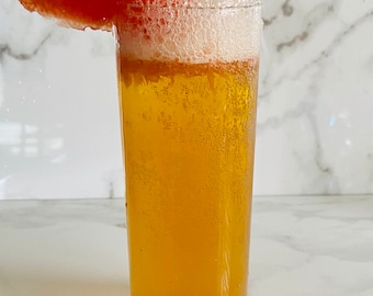 May include: A tall glass filled with a light yellow bubbly beverage topped with a thick layer of white foam. A slice of grapefruit is perched on the rim of the glass.