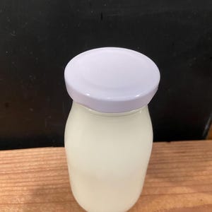 May include: A clear glass jar filled with a white liquid, topped with a white lid. The jar sits on a wooden surface against a dark background. The jar has a rounded shape.