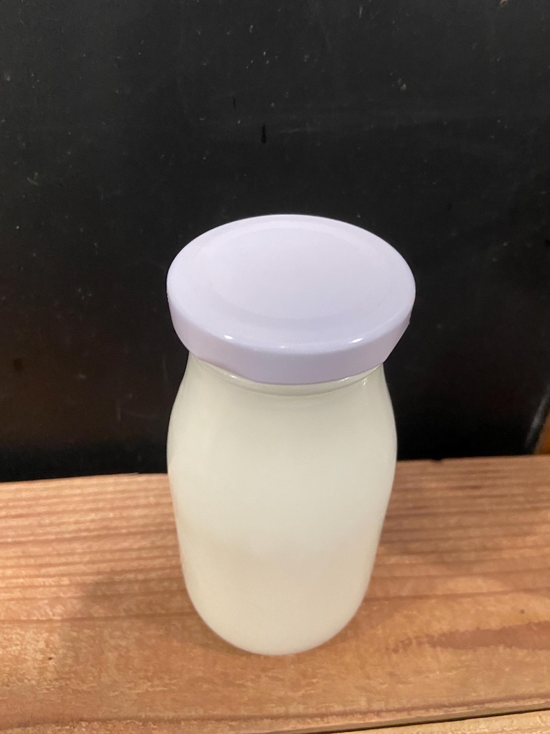 May include: A clear glass jar filled with a white liquid, topped with a white lid. The jar sits on a wooden surface against a dark background. The jar has a rounded shape.